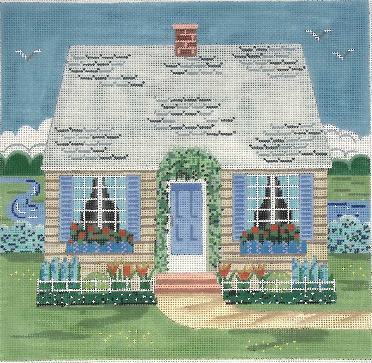 A cross stitch kit of a cottage with blue shutters and flowers.